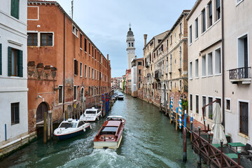 Fototapeta na wymiar MAY 20, 2017 - VENICE, ITALY: Water taxi crossing picturesque canal surrounded by vintage buildings, with bridge and bell tower in the distance.