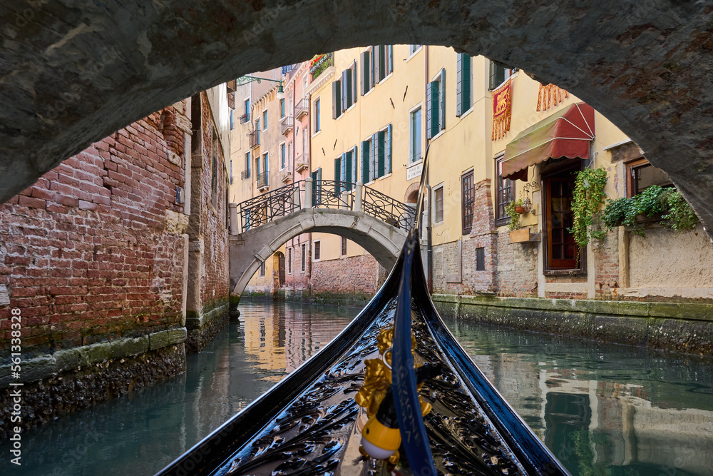 Wall mural Gondola ride going under a bridge in Venice, view of canal and old bridge surrounded by picturesque buildings, details of brick walls and reflections on the water. Romantic view from gondola.