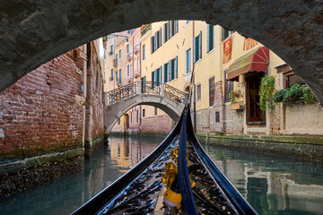 Gondola ride going under a bridge in Venice, view of canal and old bridge surrounded by picturesque buildings, details of brick walls and reflections on the water. Romantic view from gondola. © Eduardo Accorinti