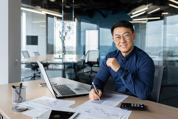 Obraz premium A successful young man businessman Asian works in the office at the table, works with documents, graphs, plan, accounts. Writes, looks at the camera, smiles.