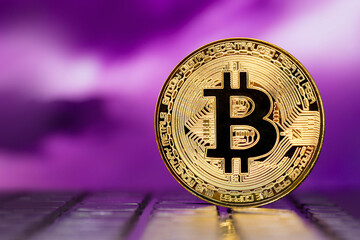 Golden metallic Bitcoin coin standing on a laptop keyboard against the background of a blurred  multicolored sky 