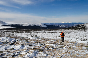 Obraz premium Mountain winter landscape. A tourist with a backpack walks through a snow-covered field. In the background, snowy mountains, blue sky.