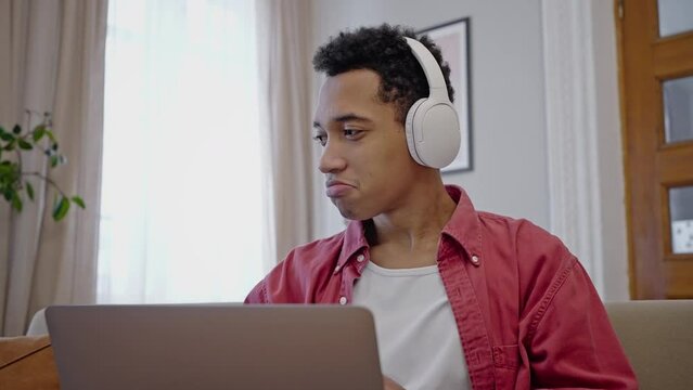 Charming African American young man working on laptop and moving listening to music on headphones