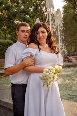 Woman in lush white dress poses against the backdrop of afountain in park, man in white shirt stands behind