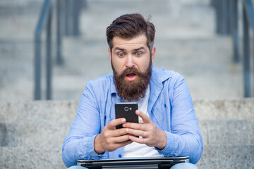 Surprised man checking up mobile phone betting results while sitting on steps outdoors