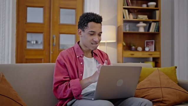 Handsome young African American man, freelancer types a text on laptop, sitting on the sofa at home