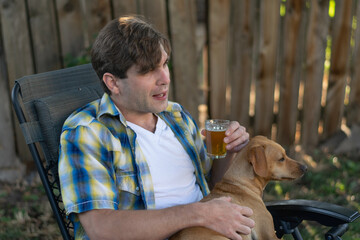 A young white with a dog in his lap man sips a beer outdoors in a shady backyard setting. 