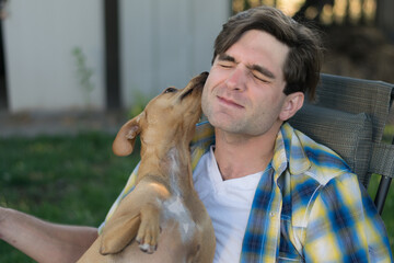 A dog gives a kiss to a young white man in a shady outdoor setting.