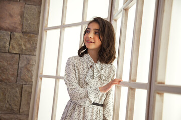 Portrait pretty girl in stylish white dress posing near large light window in living room, turned around. Studio shot positive teenager lady. Child emotion concept. Copy text space for advertising