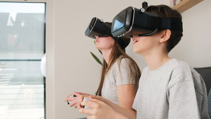 Caucasian girl and her young brother having fun at home, playing driving simulation video games by using virtual reality headsets and controllers