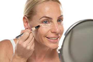 Middle aged senior woman applyes concealer under her eyes on white background