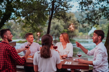 Group of happy friends celebrating holiday vacation using sprinklers and drinking red wine while having picnic french dinner party outdoor near the river on beautiful summer evening in nature