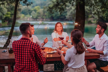Group of happy friends celebrating holiday vacation using sprinklers and drinking red wine while having picnic french dinner party outdoor near the river on beautiful summer evening in nature