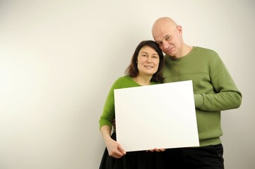 couple holding banner adult modest man and woman in green clothes hold poster in front of them on white background with no inscriptions look at each other woman points finger place for text of ad