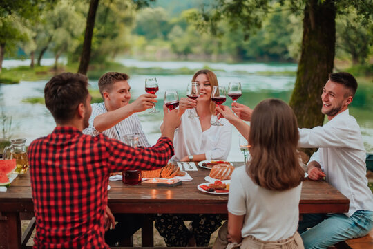 Group Of Happy Friends Toasting Red Wine Glass While Having Picnic French Dinner Party Outdoor During Summer Holiday Vacation Near The River At Beautiful Nature