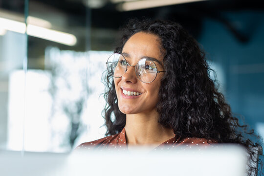 Portrait of successful businesswoman wearing glasses inside office, woman looking out window smiling and dreaming happy with achievement results, Hispanic woman at workplace with laptop.