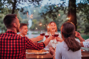 Group of happy friends celebrating holiday vacation using sprinklers and drinking red wine while having picnic french dinner party outdoor near the river on beautiful summer evening in nature