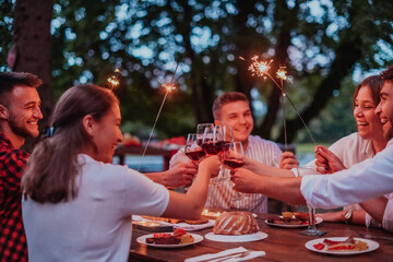 Group of happy friends celebrating holiday vacation using sprinklers and drinking red wine while having picnic french dinner party outdoor near the river on beautiful summer evening in nature