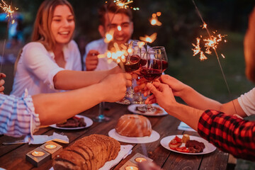 Group of happy friends celebrating holiday vacation using sprinklers and drinking red wine while having picnic french dinner party outdoor near the river on beautiful summer evening in nature