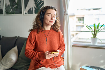 Calm mindful pretty young woman wearing headphones using mobile phone listening music, audio...