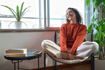 Fotobehang Young relaxed smiling pretty woman relaxing sitting on chair at home. Happy positive beautiful lady feeling joy enjoying wellbeing and lounge chilling near window in modern cozy apartment interior. © insta_photos