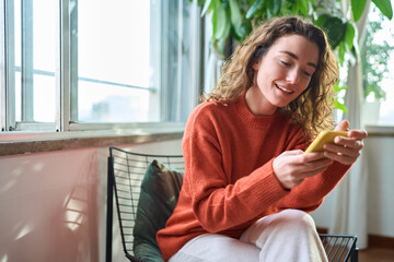 Smiling young woman sitting on chair holding mobile phone using cellphone device, looking at smartphone, checking modern apps, texting messages, browsing internet doing shopping relaxing at home. - obrazy, fototapety, plakaty