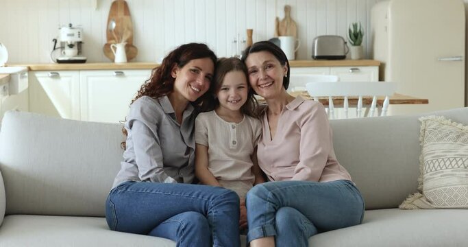 Beautiful multi generational family portrait. Older woman her grown up daughter and little granddaughter sit on sofa at home shooting for family photo, enjoy good harmonic relations and time together