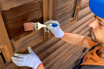 Worker applying protective varnish or wood oil on wooden house cottage exterior walls and window...