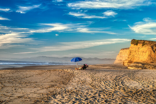 Two People In Distance Sitting Under An Umbrella At The Beach