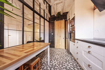 Furnished kitchen of a vacation rental loft with a French-style black metal partition, long wooden table, appliance column and hydraulic stoneware floor