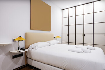 Bedroom with a double bed with a white feather duvet with white shelves as bedside tables with twin lamps and a large built-in wardrobe with white wooden doors with bars