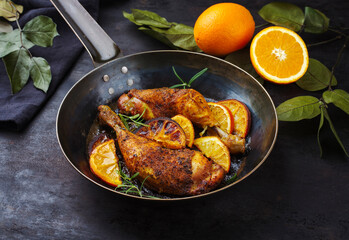 Traditional barbecue chicken drumsticks with orange slices served with herbs as close-up on a rustic metal frying pan