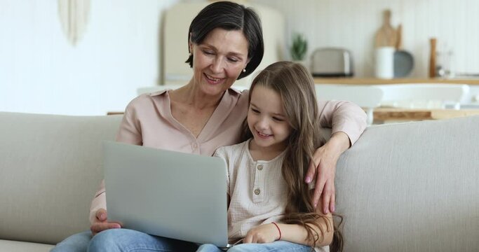 Older granny sits on sofa with granddaughter use laptop, discuss new vlog content watch online movie, cartoons, spend carefree leisure together at home. Child development use internet, family pastime