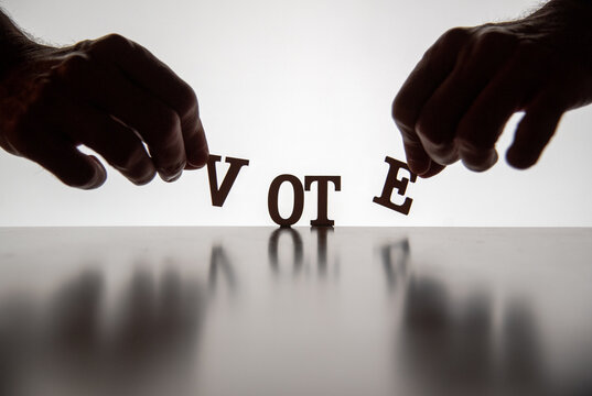 Silhouetted Hands Placing The Letters For The Word VOTE On Reflective Surface Backlit By White. 