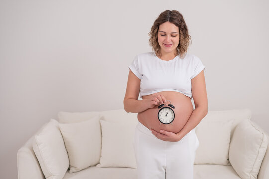 A pregnant woman with a naked belly holds an alarm clock. Intended date of birth.