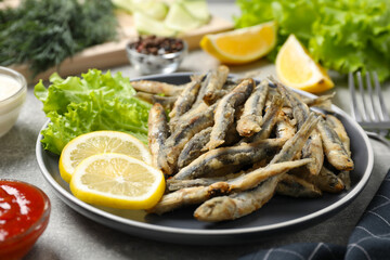 Plate with delicious fried anchovies, lemon, lettuce leaves and sauces served on light grey table, closeup