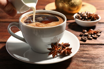 Woman pouring milk into cup of coffee with anise stars at wooden table