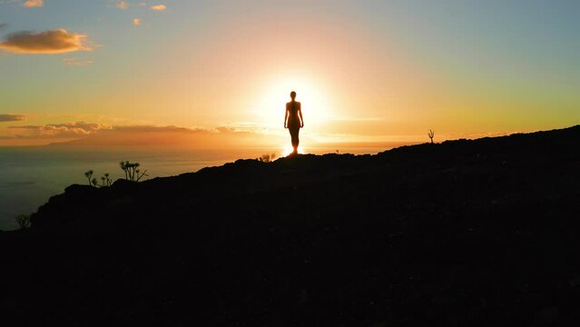 Lonely woman silhouette on mountain top during orange sunset drone flight. Sun rays otline a person. Hiker enjoying sunrise. Sunbeams rise from ocean.