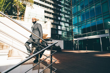 A black white-collar employee moving up the staircase, a metallic railing next to him, the office building with blue windows behind him and he is carrying a weekender bag, wearing dark grey suit