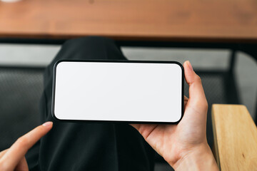 Hands holding smartphone with mockup of blank screen on sofa in living room.