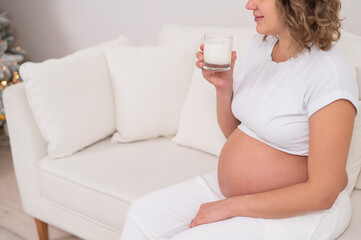 A pregnant woman sits on a white sofa and drinks milk.