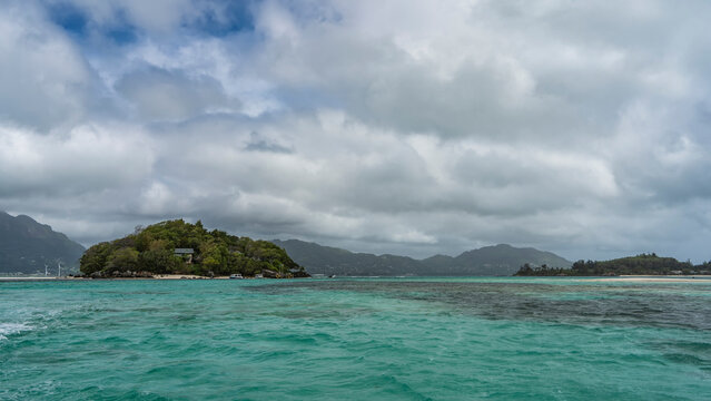 Small islands in the turquoise ocean are overgrown with tropical vegetation. Cottages are visible through the foliage of trees. The boats are moored at the shore. Clouds in the sky. Seychelles