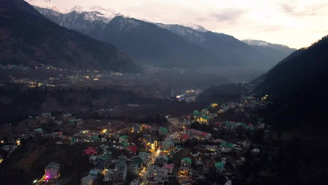 aerial drone shot flying forward of hill station town manali shimla in valley with lights on buildings with fog covered himalaya mountains in distance shot in evening himachal pradesh India