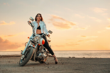 Fototapeta na wymiar Sexy woman with a high heels and leather pants, posing with motorcycle. Sunset sky on the background. Copy space. The concept of Motorcyclist Day