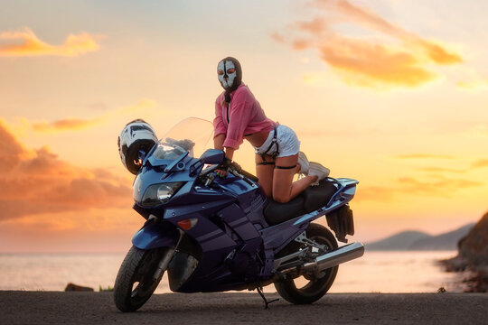 Sexy woman in a balaclava with skull poses on a tourist motorcycle. Beautiful golden sunset in the background. The concept of moto travel and motorcycle world day