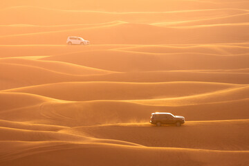 Two offroad 4x4 are racing over the ridge of dunes in a sandy landscape during the golden hour