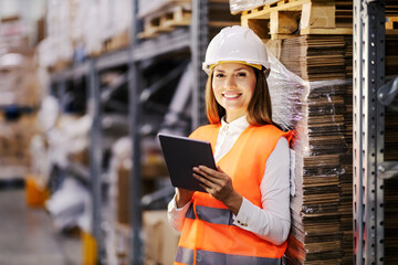 Portrait of a happy businesswoman leaning on shelf in storage and tracking shipment on tablet while...