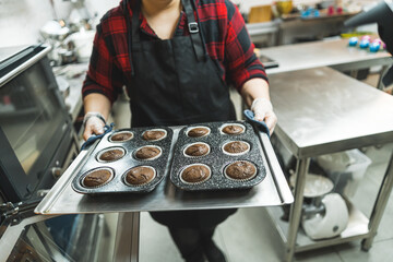 Female baker wearing black apron walking to the oven to bake tray of chocolate cupcakes. Professional baking. Horizontal indoor shot . High quality photo