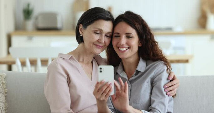 Adult Hispanic daughter and older 50s mother rest on sofa, holding smartphone, watch photos and videos, recollects, enjoy memories spend leisure together at home using gadget. Modern tech, e-commerce