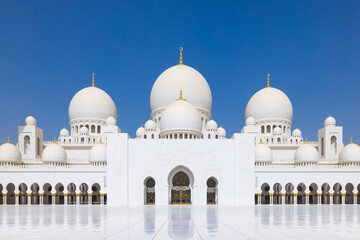 Symmetrical close up of the main courtyard of the Sheik Zayed grand mosque in Abu Dhabi with the prayer hall and its round domes against a blue sky